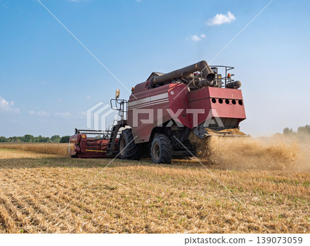 A combine harvester working in a vast agricultural field during a sunny daytime harvest, kicking up dust A combine harvester working in a vast agricultural field during a sunny daytime harvest, kicking up dust 139073059
