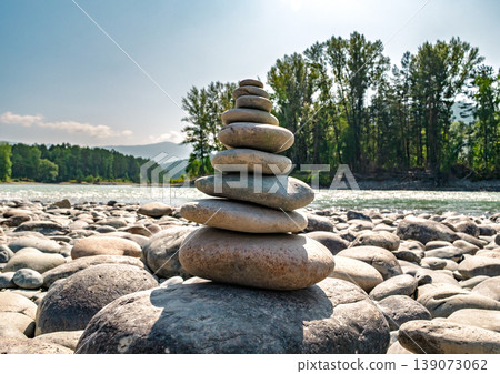 Smooth river stones creating a balanced cairn on a sunny riverbank, with a flowing river and green forest Smooth river stones creating a balanced cairn on a sunny riverbank, with a flowing river and green forest 139073062