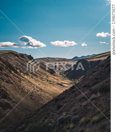 Sunlit valley expanse in the Altai Mountains with rugged slopes and a clear blue sky dotted with white clouds Sunlit valley expanse in the Altai Mountains with rugged slopes and a clear blue sky dotted with white clouds 139073077