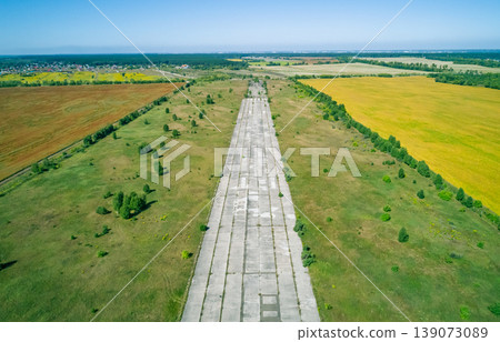 Aerial view of a long, cracked concrete runway at an abandoned airfield, flanked by green fields and dense trees. Aerial view of a long, cracked concrete runway at an abandoned airfield, flanked by green fields and dense trees. 139073089