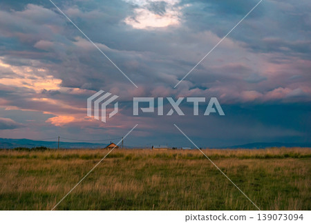 Dramatic clouds hover above a solitary rural house in a vast grassy field at twilight Dramatic clouds hover above a solitary rural house in a vast grassy field at twilight 139073094