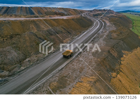 Aerial view shows large dump truck transporting materials on a wide dirt road at an open pit mine site during daytime 139073126