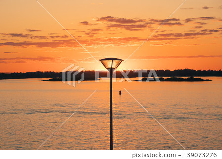 Street lamp silhouetted against a vibrant orange and yellow sky during a beautiful sunset over a wide river Street lamp silhouetted against a vibrant orange and yellow sky during a beautiful sunset over a wide river 139073276