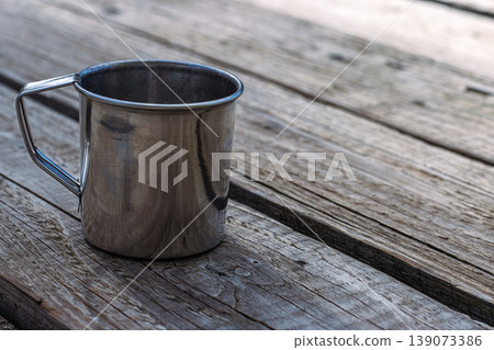 Stainless steel mug on rustic wooden table outdoors, reflecting sunlight. Metal coffee cup. Minimalism, travel essentials, camping, hiking, sustainable living in nature. Selective focus Stainless steel mug on rustic wooden table outdoors, reflecting sunlight. Metal coffee cup. Minimalism, travel essentials, camping, hiking, sustainable living in nature. Selective focus 139073386