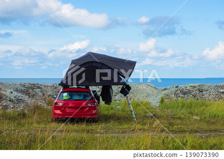 Red car with a rooftop tent parked near the sea, perfect for camping, adventure, and road trip lifestyle in nature. Outdoor exploration, freedom concept. Blue cloudy sky on background. Copy space 139073390