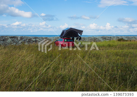 Red car with a rooftop tent parked near the sea, perfect for camping, adventure, and road trip lifestyle in nature. Outdoor exploration, freedom concept. Blue cloudy sky on background. Copy space 139073393