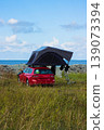 Red car with a rooftop tent parked near the sea, perfect for camping, adventure, and road trip lifestyle in nature. Outdoor exploration, freedom concept. Blue cloudy sky on background. Copy space 139073394