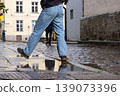 Person in blue jeans stepping over a puddle on a wet cobblestone street after rain, reflecting city architecture and urban lifestyle in Tallinn, Estonia, old town. 139073396