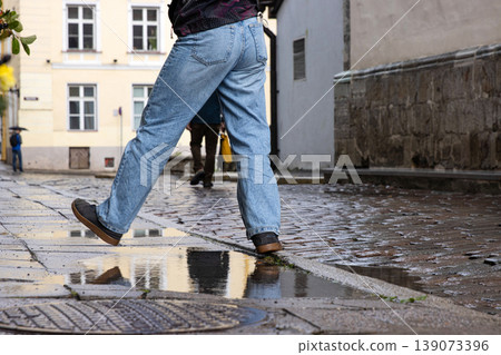 Person in blue jeans stepping over a puddle on a wet cobblestone street after rain, reflecting city architecture and urban lifestyle in Tallinn, Estonia, old town. 139073396