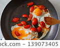 Overhead view of two fried eggs cooked with cherry tomato pieces in a dark frying pan, with a wooden spatula, Simple homemade breakfast. Cooking scene 139073405