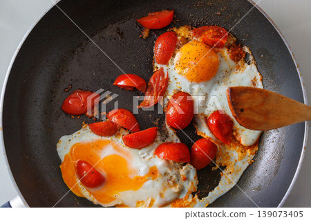Overhead view of two fried eggs cooked with cherry tomato pieces in a dark frying pan, with a wooden spatula, Simple homemade breakfast. Cooking scene 139073405
