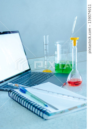 Laboratory workstation showing scientific research setup with a laptop, notebook, pen, ruler, and various glass beakers and flasks containing colorful liquids Laboratory workstation showing scientific research setup with a laptop, notebook, pen, ruler, and various glass beakers and flasks containing colorful liquids 139074011
