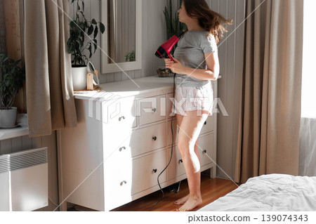 A woman blow-drying her hair, soft morning sunlight shining through the curtains, a concentrated expression on her face, hands running through chestnut curls, a home routine, relaxed self-care. A woman blow-drying her hair, soft morning sunlight shining through the curtains, a concentrated expression on her face, hands running through chestnut curls, a home routine, relaxed self-care. 139074343