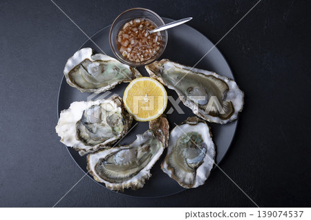 Khasan oysters, live, fresh, opened. Served with classic French Mignonette sauce and half a lemon. Blue plate, black background. Selective focus, close-up. Khasan oysters, live, fresh, opened. Served with classic French Mignonette sauce and half a lemon. Blue plate, black background. Selective focus, close-up. 139074537