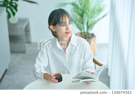 A woman reading a book by the window in her room. 139074561