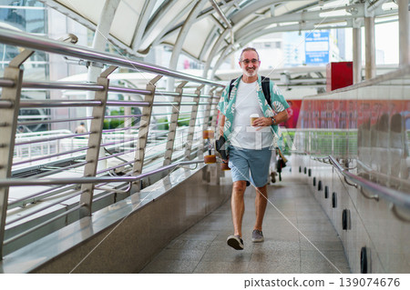 Happy senior Caucasian man with white beard and sunglasses walking on city skywalk while carrying electric skateboard. Concept of joyful aging, travel adventure, and the use of modern personal mobilit Happy senior Caucasian man with white beard and sunglasses walking on city skywalk while carrying electric skateboard. Concept of joyful aging, travel adventure, and the use of modern personal mobilit 139074676