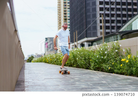 Cheerful senior Caucasian man with beard and glasses rides longboard along Skywalk in downtown district. Focusing on active retirement lifestyle and urban tourism. Concept of joyful aging and vacation Cheerful senior Caucasian man with beard and glasses rides longboard along Skywalk in downtown district. Focusing on active retirement lifestyle and urban tourism. Concept of joyful aging and vacation 139074710