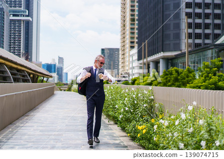 Professional senior Caucasian businessman in navy vest and tie, holding a coffee cup and carrying a backpack. He is looking up at urban skyscrapers in modern business district. Portrait of senior male 139074715