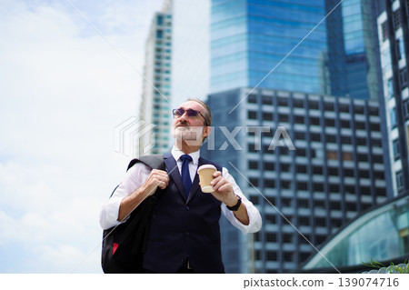 Professional senior Caucasian businessman in navy vest and tie, holding a coffee cup and carrying a backpack. He is looking up at urban skyscrapers in modern business district. Portrait of senior male Professional senior Caucasian businessman in navy vest and tie, holding a coffee cup and carrying a backpack. He is looking up at urban skyscrapers in modern business district. Portrait of senior male 139074716