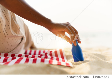 Close up shot of woman hand reaching for blue sunscreen bottle placed on red and white striped towel over the sand. Perfect for summer skincare, sun protection, and tropical vacation themes. Close up shot of woman hand reaching for blue sunscreen bottle placed on red and white striped towel over the sand. Perfect for summer skincare, sun protection, and tropical vacation themes. 139074837