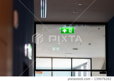 A green fire exit sign hangs from the ceiling with a running man and right arrow, combining safety direction with emergency evacuation guidance in an indoor building. A green fire exit sign hangs from the ceiling with a running man and right arrow, combining safety direction with emergency evacuation guidance in an indoor building. 139076262