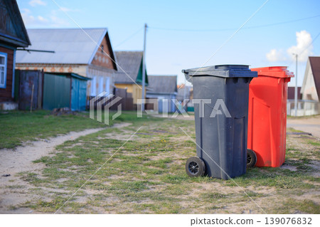 Pair of plastic garbage bins for selective waste disposal on a village street. Black and red recycling containers in a rural neighborhood. 139076832