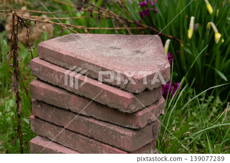 Stack of weathered red pavers in garden surrounded by green grass and purple flowers with yellow buds. Stack of weathered red pavers in garden surrounded by green grass and purple flowers with yellow buds. 139077289