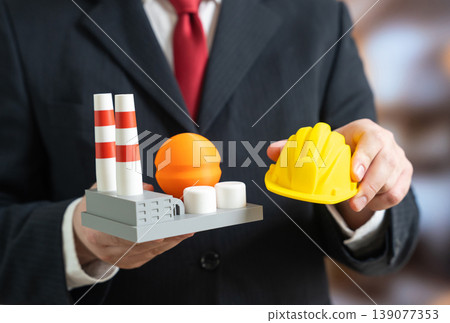 A factory and a construction helmet in the hands of a businessman. Building a new industrial plant supports modernization. Energy upgrades improve efficiency and production capacity. A factory and a construction helmet in the hands of a businessman. Building a new industrial plant supports modernization. Energy upgrades improve efficiency and production capacity. 139077353