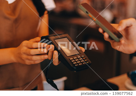 Asian senior female barista adult receiving mobile payment from customer using smartphone at cafe demonstrating modern cashless transaction in cozy local family coffee shop 139077562