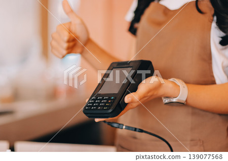 Friendly Asian female barista receives a credit card from a customer for payment. Both women are smiling in a warmly lit coffee shop with a rustic interior. Cashless payments. Small family business 139077568