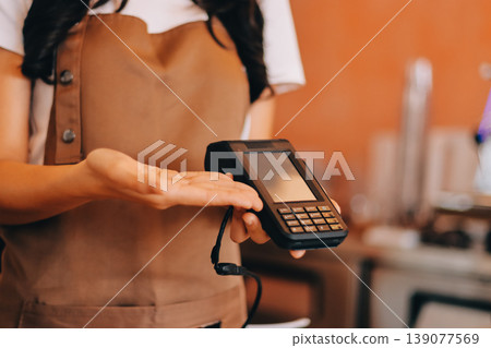 Friendly Asian female barista receives a credit card from a customer for payment. Both women are smiling in a warmly lit coffee shop with a rustic interior. Cashless payments. Small family business 139077569