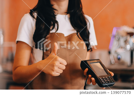Friendly Asian female barista receives a credit card from a customer for payment. Both women are smiling in a warmly lit coffee shop with a rustic interior. Cashless payments. Small family business 139077570