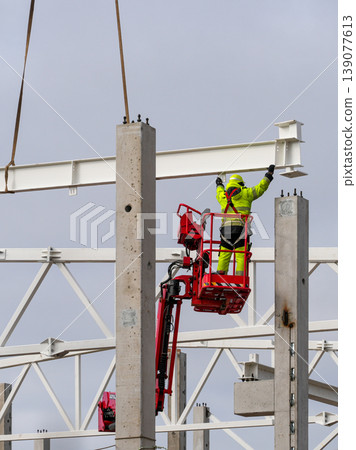 Construction worker guiding steel beam into position on concrete columns using crane and lift Construction worker guiding steel beam into position on concrete columns using crane and lift 139077613