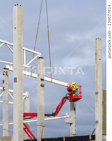 Worker in aerial lift guiding steel beam during industrial building frame construction project Worker in aerial lift guiding steel beam during industrial building frame construction project 139077614