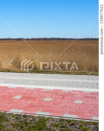 Red bicycle lane and sidewalk at edge of rural field with tall grass under clear blue sky Red bicycle lane and sidewalk at edge of rural field with tall grass under clear blue sky 139077622