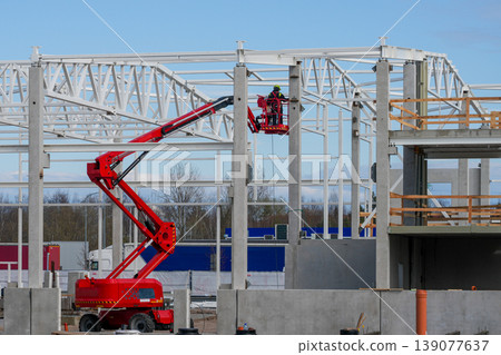 Worker on red boom lift installing steel frame on industrial building construction site 139077637