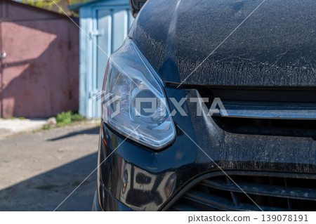 A black car is parked on the street. The light on the front is visible. A blue container is in the background A black car is parked on the street. The light on the front is visible. A blue container is in the background 139078191