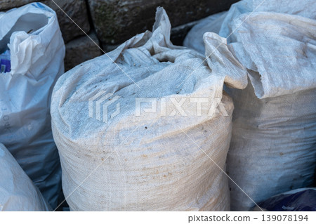 Several large bags filled with waste materials are stacked on the ground next to a stone wall in a non-urban area during the day Several large bags filled with waste materials are stacked on the ground next to a stone wall in a non-urban area during the day 139078194