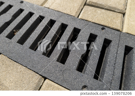 A drain cover sits on gray paving stones under bright sunlight on a clear day. The drain features several slits and holes 139078206