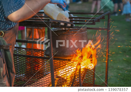 Large pot is cooking over an open fire, cook pours ingredients into the pot during outdoor food preparation. Cooking over fire, traditional cuisine, rustic food preparation 139078357