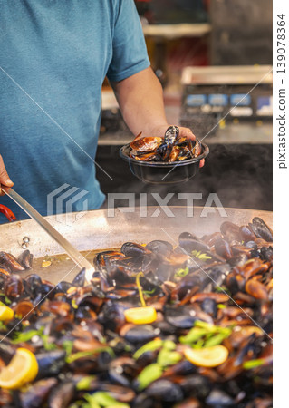 Fresh mussels are being served hot from large pan at bustling street food stall, steam rising, vibrant ingredients visible. Mussels cooking, street cuisine culture, and fresh seafood preparation Fresh mussels are being served hot from large pan at bustling street food stall, steam rising, vibrant ingredients visible. Mussels cooking, street cuisine culture, and fresh seafood preparation 139078364
