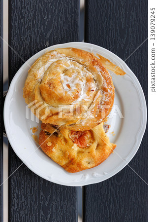 Top view of a white plate with a sweet burek pastry roll and a piece of focaccia with olive and tomato on a dark wooden table with copy space. Top view of a white plate with a sweet burek pastry roll and a piece of focaccia with olive and tomato on a dark wooden table with copy space. 139079245