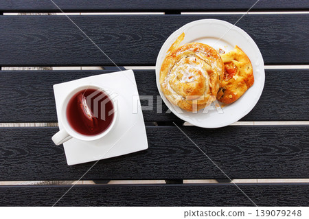 Overhead view of a cup of red fruit tea and a white plate with sweet burek pastry roll and focaccia on a dark slatted balcony table. Copy space 139079248