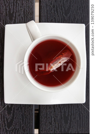 Overhead view of a cup of red fruit tea with a teabag in a white cup on a square white saucer, placed on a dark slatted table. Copy space. Selective focus Overhead view of a cup of red fruit tea with a teabag in a white cup on a square white saucer, placed on a dark slatted table. Copy space. Selective focus 139079250