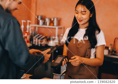 Asian senior female barista adult receiving mobile payment from customer using smartphone at cafe demonstrating modern cashless transaction in cozy local family coffee shop 139082820