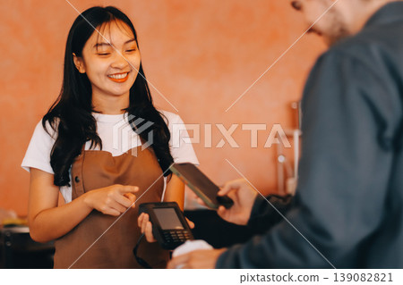 Asian senior female barista adult receiving mobile payment from customer using smartphone at cafe demonstrating modern cashless transaction in cozy local family coffee shop 139082821