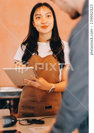 Friendly Asian female barista receives a credit card from a customer for payment. Both women are smiling in a warmly lit coffee shop with a rustic interior. Cashless payments. Small family business 139082823