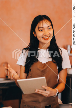 Friendly Asian female barista receives a credit card from a customer for payment. Both women are smiling in a warmly lit coffee shop with a rustic interior. Cashless payments. Small family business 139082826