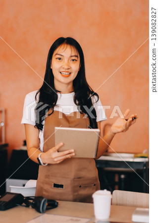 Friendly Asian female barista receives a credit card from a customer for payment. Both women are smiling in a warmly lit coffee shop with a rustic interior. Cashless payments. Small family business 139082827