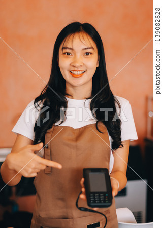 Friendly Asian female barista receives a credit card from a customer for payment. Both women are smiling in a warmly lit coffee shop with a rustic interior. Cashless payments. Small family business 139082828